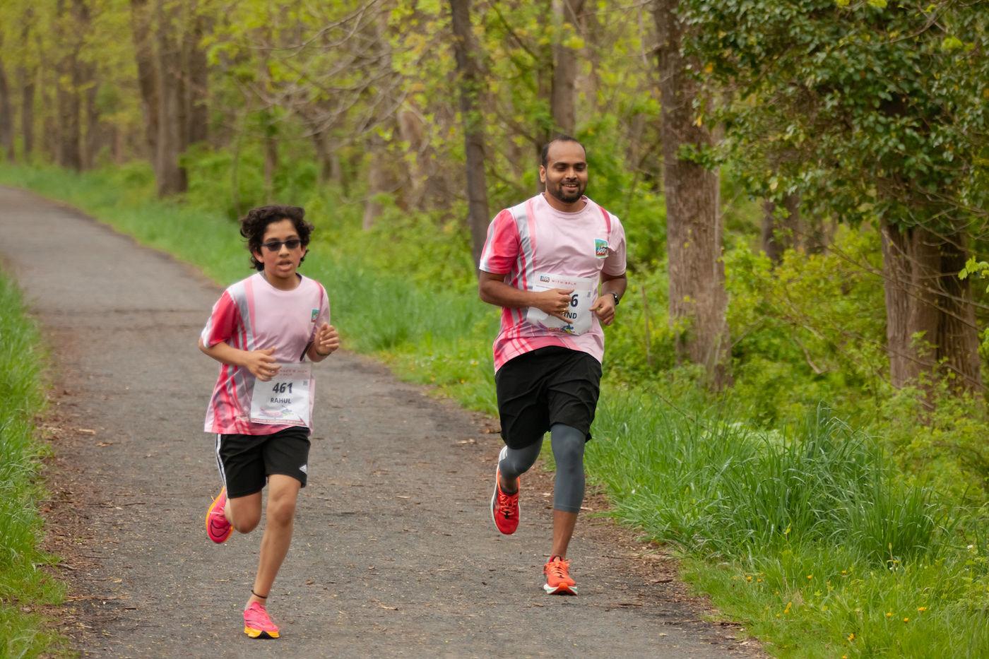 A young runner and an adult running together on a wooded trail